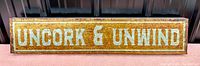 Front view of the rectangular metal sign with the phrase 'Uncork & Unwind' in white capital letters on a rustic brown background.