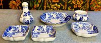 Front and angled view of all six Royal Crown Derby Mikado pattern pieces on a blue table surface: salt shaker, two butter pats, two trinket dishes, one bud vase, showing blue and white scenic designs and gold trim.