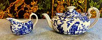 Front view of teapot and creamer side by side on gray surface with tapestry background
