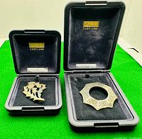 Two sterling silver brooches in their original black velvet-lined Ortak Scotland boxes against a green background.