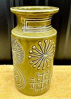 Front angled view showing the olive green ceramic vase with prominent raised totemic patterns including starburst and geometric motifs.