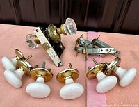 Photo shows four white porcelain oval door knobs with brass bases, the glass knob on the black metal lock mechanism, and assorted metal lock parts arranged on a pink cloth.