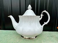 Side view of large white bone china Royal Albert Chantilly teapot with curved spout, ornate handle and finial on lid.