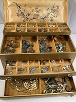 Photo of four drawers of costume jewelry displayed in an open gold fabric-lined jewelry box showing various earrings, bracelets, necklaces, and pins arranged in compartments.