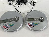 Two Sony silver circular CD Walkman D-NF340 players side-by-side with tangled black wired earbuds.