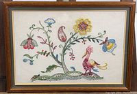 Full view of framed crewel embroidery showing bird with flower motif and wood frame