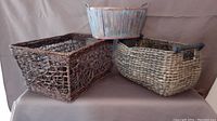 Three baskets displayed side by side with neutral fabric background. One is twig-like weave rectangular, second is elongated woven with dark handles, third is round wooden bucket style with metal handles.