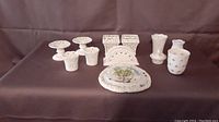 Photo showing assortment of 11 white porcelain knickknack dishes and decorative pieces arranged on dark brown fabric. Pieces include small pedestal dishes, salt and pepper shakers, napkin holders, vases, and a floral decorated tray.