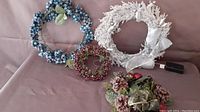 Photo showing four wreaths: white lighted battery-operated wreath, blue berry wreath, small red berry wreath, pinecone and ornament wreath.