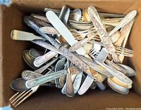 Top view of a box filled with assorted vintage forks, spoons, and butter knives showing tarnish and rust spots.