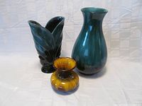 Three Blue Mountain Pottery vases displayed on white cloth background: tall dark green vase, leaf-shaped dark green vase, small harvest gold vase.