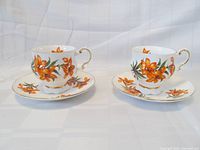 Front view showing two teacup and saucer sets with orange prairie lily floral pattern and gold trim around rims and handles.