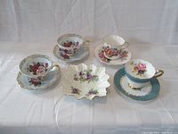 Four floral fine bone china teacup and saucer sets with varying floral patterns and one square mint dish with purple flowers and fluted edges, all on white background.