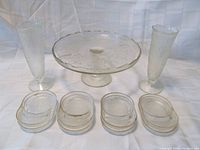 Eight clear glass coaster/ashtrays arranged in two rows in front, two slender 7.5 inch vases, and one 9 inch footed cake plate behind them on a white fabric background
