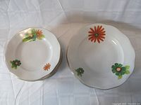 Stack of 12 bread and butter plates on left, 4 larger dinner plates on right. Plates are white porcelain with orange and green flower decorations and gold trim.