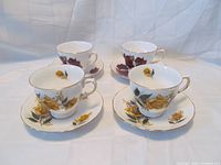 Four total teacup and saucer sets displayed with two yellow rose pattern sets in front and two maroon leaf pattern sets in back.