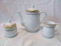 Three-piece tea set including teapot, creamer, and sugar bowl with lids, photographed together on white fabric backdrop.