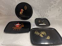 Four black lacquer holiday-themed serving trays by Couroc Monterey California on a white patterned fabric background.