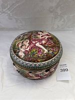 Front angle of closed circular Capodimonte porcelain box showing colorful raised relief figures and floral pattern on lid and sides.