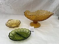 Three vintage glass bowls: large amber pedestal bowl, small amber bowl, and green divided dish on white fabric background.