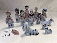 Front view of eleven miniature Mexican ceramic nativity figurines arranged on a table with some figures standing and others lying down, showing human and animal figures with pastel pink and blue glazing and black painted details.