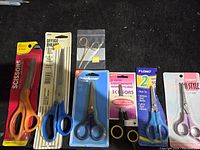 Overview of 7 pairs of new assorted scissors laid out showing varying handle colors and packaging styles