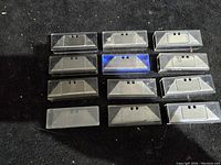 Photo showing rows of trapezoidal razor blades in mostly transparent plastic protective cases, blades appear new and unused