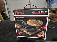 Box front showing the Parini 2-liter cast iron noodle hot pot with wooden lid and trivet, emphasizing its Asian Fusion style and usage for noodles, soups and more.