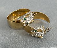 Photo showing two gold plated earrings with cat-shaped heads featuring green stones and clear stones on the face.