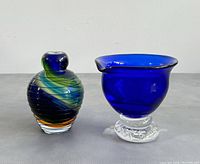 Two handblown glass pieces: a small vase with multicolored swirling stripes and a cobalt blue pedestal bowl with clear textured base, shown side by side on a gray surface