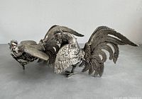 Pair of silver-coloured cast metal roosters on flat surface, showing detailed feather textures and posture.