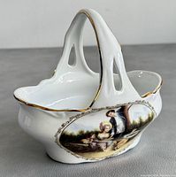 Side view of the porcelain basket showing the painted classical scene and gold trim on the scalloped edges.