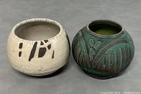 Two raku pottery bowls side by side on grey surface. Left bowl is cream-colored with black markings and natural craquelure. Right bowl is dark green with carved patterns and black rim.