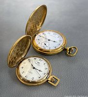 Two open watches showing white enamel dials, Arabic numerals and hands.
