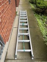 Ladder in collapsed position showing entire length on concrete pathway beside brick wall, secured with green rope.