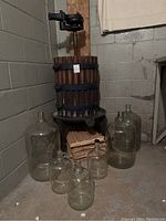 Photo of wooden grape crusher press with metal bands and black metal crank handle mounted on metal stand, surrounded by seven clear glass carboys of different sizes and stacked wooden slatted crates beneath.