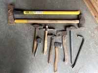 Top-down view of 10 heavy hand tools arranged on concrete floor including 2 sledgehammers, multiple hammers, a rubber mallet, hatchets, and crowbar.
