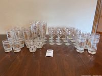 Full lot of assorted vintage Libbey glasses in various sizes with frosted white backgrounds and gold gilt pinecone designs, arranged on wood table.