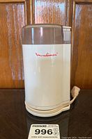 Front view of white Moulinex electric coffee grinder with logo and power cord wrapped around base, taken against wood background.