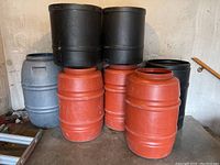 Group shot of six rain barrels in various colors including terra cotta, black, and gray, stacked and arranged on a concrete floor.