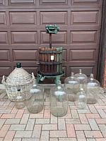 Full view of wine making equipment including wooden crusher, green metal stand, multiple glass carboys including one in wicker basket, on outdoor brick floor.