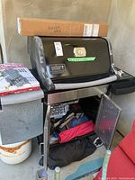 Photo showing Weber Genesis Silver gas BBQ with burner tube parts in a box on top, open storage cabinet with utensils and accessories inside, propane tank below.