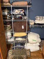Closet with wire shelf holding stacked bed linens, blankets, and pillows. Two patterned rugs rolled and placed on the wooden floor in front.