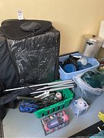 Overview of camping items including foam folding bed, camping plates and pans in blue plastic container, camping chairs folded, and various small camping accessories in green crate.