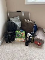 Photo of camping supplies including gray and red Outbound Eiger 4 tent carry bag, brown sleeping bag, binoculars, Coleman cooler, thermos, travel mug and other gear.