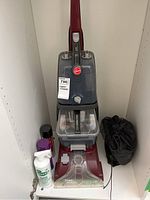 Full front view of Hoover Power Scrub carpet cleaner in red and gray with water tanks, control knob, and cleaning brush base visible. Several cleaning sprays and a black storage pouch placed around the cleaner on floor and shelf.
