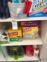 Shelf with various cleaning products and garbage bags including Glad compost and kitchen bags, Kirkland kitchen bags, OxiClean, plastic bins, and metal bucket with cloth.