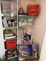 Shelf unit holding multiple Christmas decorative items including boxed fibre optic wreath and Santa's house, stacked boxes of tree ornaments, lights, mugs and wall pictures.