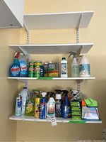 Household cleaning supplies arranged on white wall shelves showing various spray bottles and liquid cleaners including Windex, Comet, Lysol, Vim, and others