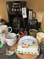 Overall lot image showing Cuisinart coffee maker, coffee grinder, ceramic mugs, napkin holder, and trivets on countertop